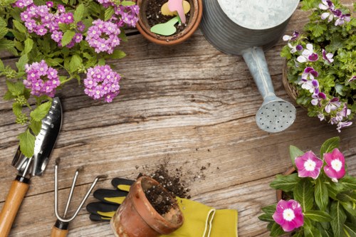The image depicts a section of a garden or outdoor planting area with a weathered wooden surface, possibly a garden table or deck, surrounded by various gardening tools and potted plants. In the top left corner, there are clusters of pink and purple flowering plants, while on the right side, similar flowering plants with bright pink flowers and green leaves are visible. In the center, a small terracotta pot filled with soil and gardening gloves placed nearby suggests ongoing planting activity. To the right of the soil pot, a silver watering can with a rounded spout rests on the surface, indicating watering or garden maintenance. Additionally, a small hand rake with a wooden handle, a trowel with a metal blade, and a container with a few soil remnants are arranged on the surface, suggesting active gardening work typical of a well-maintained garden space. The overall scene emphasizes gardening and outdoor care activities in a natural, residential setting, consistent with landscaping services provided by Landscapers Wimbledon, particularly in SW19 or surrounding areas in Wimbledon.