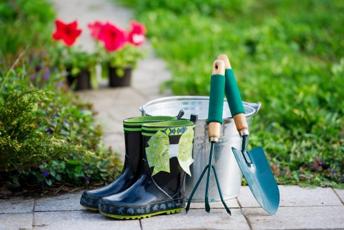 A pair of black and green rubber gardening boots with a pattern of green leaves rest on a stone patio in a well-maintained garden. Next to the boots, there are two small hand tools: a trowel and a transplanter with green handles, both leaning against a metal bucket. The background features a lush, green lawn and some potted flowering plants with red blooms, indicating recent gardening activity. The garden appears tidy and vibrant, with a combination of grass, flower beds, and paved pathways typical of a landscaped outdoor space in Wimbledon. The scene is evenly lit, suggesting a bright, mild day suitable for garden maintenance, and it highlights essential gardening tools and equipment used in outdoor landscaping and lawn care managed by companies like Landscapers Wimbledon.