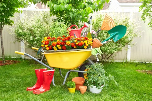 A colourful garden scene featuring a yellow wheelbarrow filled with vibrant red and yellow flowering plants, located on a well-maintained green lawn. Behind the wheelbarrow, there are garden tools including a red watering can, a green-handled scoop, an orange rake, and a teal shovel leaning against a backdrop of lush shrubs and small trees. To the side, a white pot contains a leafy green plant, with several small terracotta pots placed nearby on the grass. A pair of bright red rubber boots is positioned beside the wheelbarrow, adding a pop of colour. The garden is enclosed by a white fence, and sunlight filters through the trees, casting natural light across the scene. This outdoor space exhibits a tidy, cultivated appearance typical of a residential garden in Wimbledon, and it showcases the kind of garden maintenance and planting services offered by Landscapers Wimbledon.