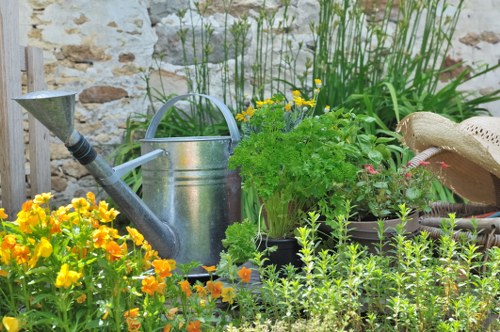 A vibrant garden scene featuring a variety of plants and gardening tools. In the foreground, there are bright yellow flowering plants alongside green foliage. Behind these, a galvanized metal watering can rests on the ground among potted plants, including a bushy green herb. To the right, part of a beige straw hat is visible, placed on top of a plant container. The backdrop showcases a rustic stone wall, adding character to the outdoor space. Tall green grasses and leafy shrubs extend upward, framing the scene and creating depth. The overall environment appears well-maintained, with natural lighting suggesting a clear day, ideal for outdoor gardening activities. This setup underscores typical garden features and tools used in lawn and plant care, aligning with services offered by companies like Landscapers Wimbledon in the Wimbledon area, with subtle references to local landscape elements.
