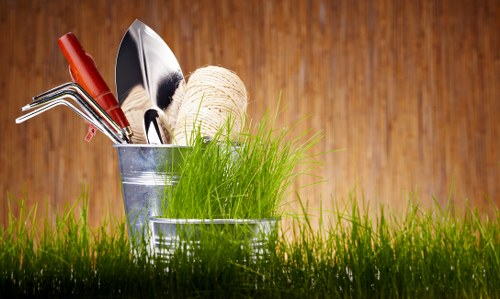 The image displays a close-up view of a small garden or outdoor space with lush, green grass in the foreground, indicating a well-maintained lawn area. Behind the grass, there are two metallic gardening buckets, one containing various hand tools including a trowel, cultivator, and pruners, along with a spool of natural twine, suggesting active gardening work. The background features a warm-toned wooden surface, which could be part of a garden shed or paneling, creating a cozy outdoor atmosphere. The scene appears to be set in natural daylight, with soft, even lighting highlighting the textures of the garden tools, grass, and wooden backdrop. This image complements gardening services offered by Landscapers Wimbledon, emphasizing maintenance, planting, and outdoor care that enhances residential gardens in Wimbledon and surrounding areas, including the local postcode region. The overall setting is neat and functional, indicating professional attention to garden organization and outdoor duty, suitable for a gardening or landscaping website dedicated to local clients seeking garden enhancement and planting services in Wimbledon.