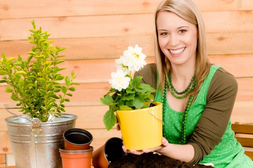 The image depicts a woman seated in a backyard or garden setting, with a wooden fence or wall in the background. In front of her are various gardening tools and pots, including a yellow pot she is holding containing a flowering plant with white blooms, and other empty pots placed nearby. To her left, there is a metallic watering can with a small green shrub inside. The woman is smiling, dressed in a green top with a beaded necklace, suggesting she is engaged in gardening or plant care activities. The outdoor space features a neatly maintained lawn area with lush, green grass covering the foreground, while the background contains a hedge or shrubbery, creating a natural, inviting garden atmosphere. The lighting indicates a bright, sunny day with clear natural light illuminating the scene, emphasizing the vibrant green tones of the plants and the cheerful expression of the gardener. This image aligns with gardening services such as plant care, pot planting, and garden upkeep that a company like Landscapers Wimbledon offers in the Wimbledon area, South West London postcode SW19.