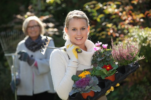 A young woman with blonde hair, wearing a white knitted sweater and gardening gloves, is standing in a well-maintained garden holding a black plant tray filled with colourful flowering plants such as pansies and chrysanthemums. Behind her, an older woman with short blonde hair, glasses, and a grey scarf around her neck is smiling while holding a gardening fork. The garden features a lush green lawn with dense shrubbery and trees in the background, some with reddish-brown leaves indicating autumn. The scene is illuminated by natural daylight, with a clear, bright atmosphere. The garden appears to be part of a landscaped outdoor space within a residential area near Wimbledon, reflecting a tidy and vibrant gardening environment suitable for professional garden and lawn care services by Landsacpers Wimbledon.