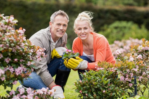 A smiling man and woman, dressed in casual outdoor clothing, are kneeling in a well-maintained garden surrounded by blooming pink and white flowering shrubs. The man has short gray hair and is wearing a light gray shirt, while the woman has blonde hair tied back and is wearing an orange cardigan. They are both wearing yellow gardening gloves and appear to be engaged in planting or tending to the flowering bushes. The garden features lush, green grass in the foreground and a background of trees and foliage, suggesting a spacious, landscaped outdoor area typical of residential gardens in Wimbledon or surrounding areas, as managed by Landscapers Wimbledon. The scene is illuminated by natural daylight, with an overall sense of a tidy, vibrant, and thoughtfully cultivated outdoor space.